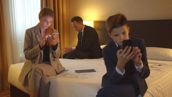 Family work on phones in hotel room, mother, father and little boy sit in suits