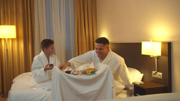 Vater und Sohn sitzen auf dem Bett im Hotelzimmer und essen das Essen — Stockfoto