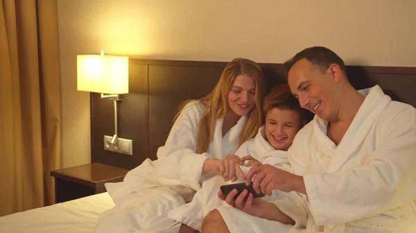 Eltern und Sohn sitzen im Bett und schauen sich Telefonfoto im Hotelzimmer an — Stockfoto