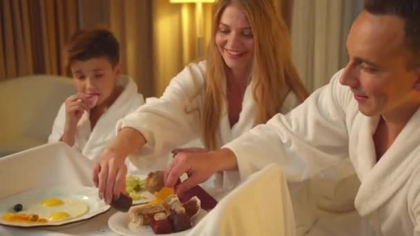 Los padres y el hijo se sientan en la cama, comen la comida y se ríen en la habitación del hotel — Vídeo de stock