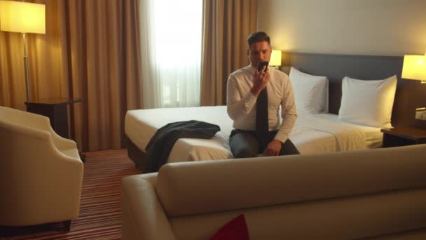 Man in shirt en stropdas zitten op het bed in het hotel en nemen voicemail op via de telefoon — Stockvideo