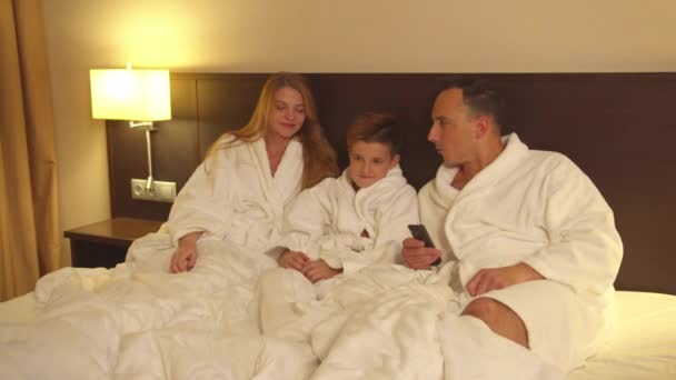 Родители и маленький сын сидят на кровати и смотрят телевизор в номере отеля — стоковое видео