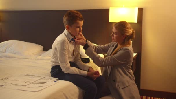 La mujer puso una corbata en el niño pequeño, que se sienta en la cama en la habitación del hotel — Vídeo de stock