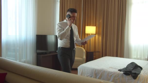 Man in shirt and tie walk in room in hotel, talk on phone and look on documents — Stock Video