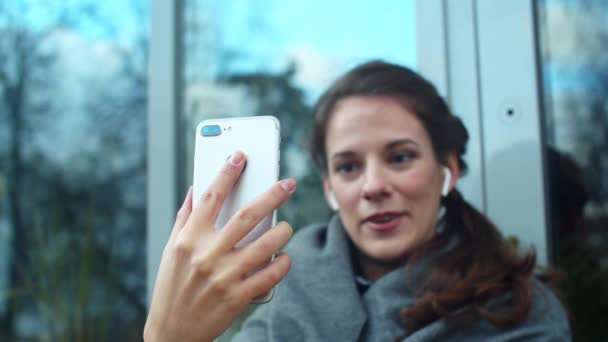 Meisje met koptelefoon zit op straat en praat aan de telefoon met een glimlach — Stockvideo
