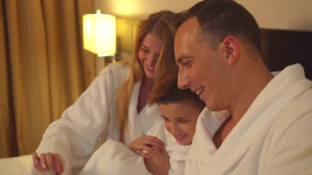 Os pais e o filho sentam-se na cama e olham para a foto no telefone no quarto do hotel — Vídeo de Stock