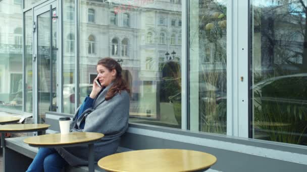 Девушка в сером одеяле сидит снаружи и разговаривает по телефону — стоковое видео