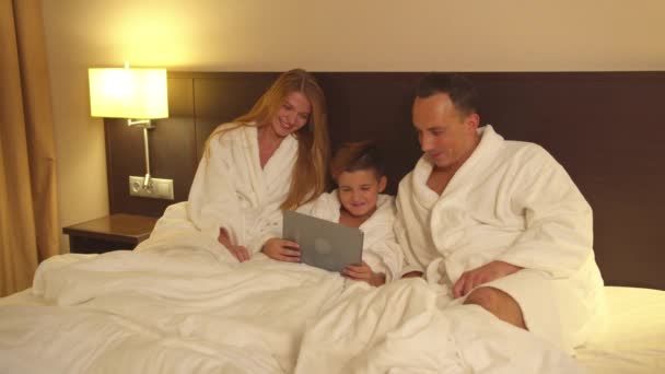 Los padres y el pequeño hijo en sentarse en la cama y ver algo en la tableta en la habitación de hotel — Vídeos de Stock