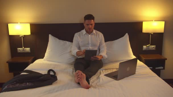 Obchodník v kalhotách sedí na posteli s tabletou v pokoji hotelu — Stock video