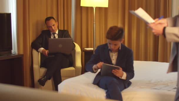 Ο άνθρωπος στην εργασία για το φορητό υπολογιστή, μικρό αγόρι κάθονται με tablet, γυναίκα ματιά σε έγγραφα στο χέρι στο δωμάτιο του ξενοδοχείου — Αρχείο Βίντεο