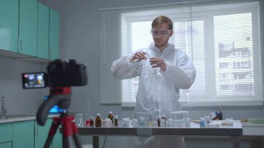 Koruyucu iş kıyafeti giyen bir adam laboratuarda kameraya sıvı döker.