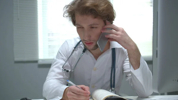 O doutor senta-se em uma mesa no armário do hospital, fala pelo telefone e escreve — Fotografia de Stock