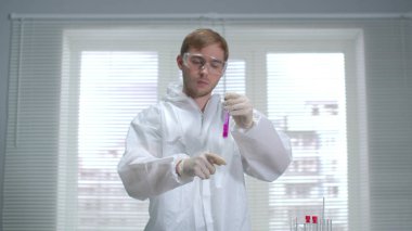 Koruyucu iş elbisesi bilim adamı tüpe bir şey döküp laboratuvarda sallıyor.
