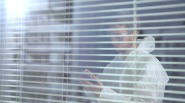 Beyaz koruyucu iş elbiseli ve gözlüklü bir adam telefonuyla laboratuvarda bekliyor.