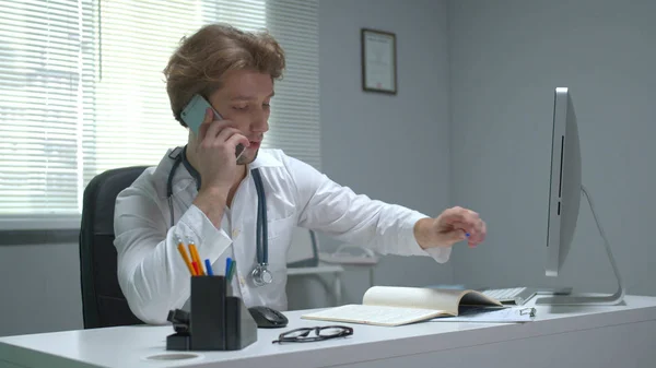 O doutor senta-se em uma mesa no armário do hospital e fala pelo telefone — Fotografia de Stock