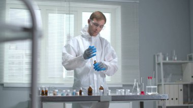 Koruyucu iş kıyafeti ve eldiven giyen adam laboratuarda kimyasal ekipmanlarla çalışıyor.
