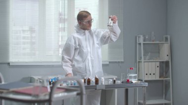 Koruyucu iş kıyafeti giyen adam laboratuarda kimyasal test tüplerini kontrol ediyor.