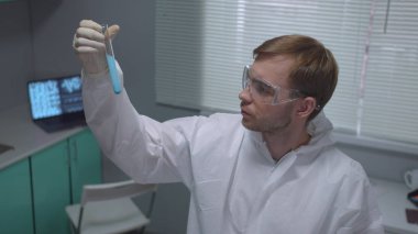 Koruyucu iş giysisindeki kimyager sıvıyla tüp yutar ve laboratuvara ciddi bir şekilde bakar.