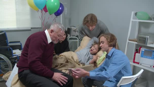 Медленное движение, родственники навещают девушку, которая лежит на кровати в больнице, мальчик показывает всем фотографии — стоковое видео