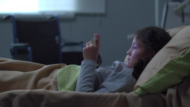 Niña acostada en la cama en el hospital, jugar con el teléfono — Vídeo de stock