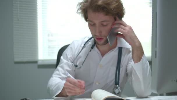 Doctor sentarse en una mesa en el gabinete del hospital, hablar por teléfono y escribir — Vídeo de stock