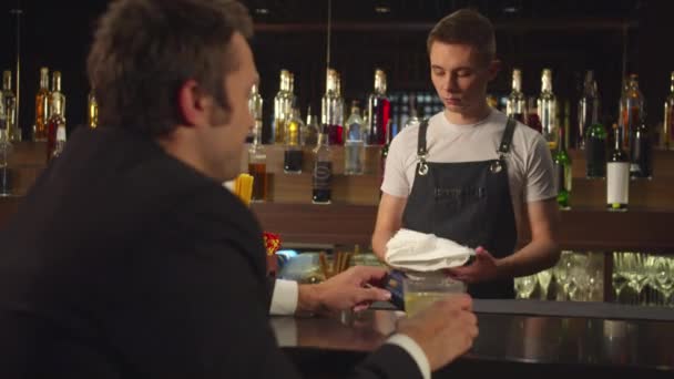 Homem senta-se no bar, tem cartão de crédito e fala com barman — Vídeo de Stock
