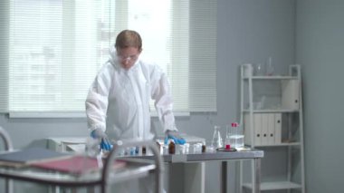 Koruyucu iş elbisesi, gözlük ve tıbbi eldiven giyen adam laboratuardaki tüpleri kontrol ediyor.