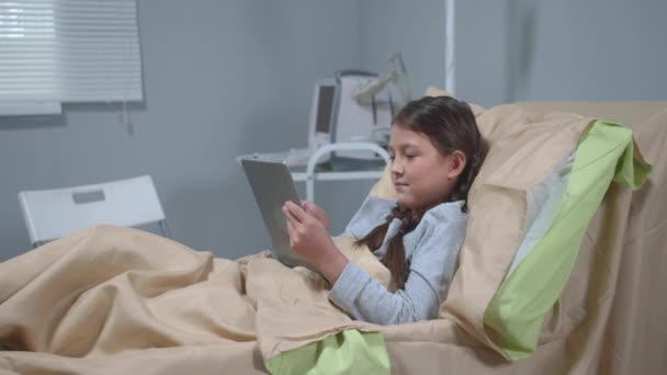 Το κορίτσι ξαπλώνει στο κρεβάτι στο νοσοκομείο και παίζει με τάμπλετ. — Αρχείο Βίντεο