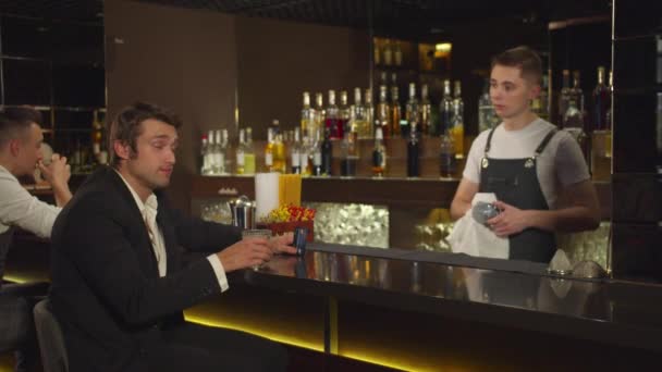 El hombre se sienta en el bar, habla con el camarero y sonríe a alguien — Vídeos de Stock