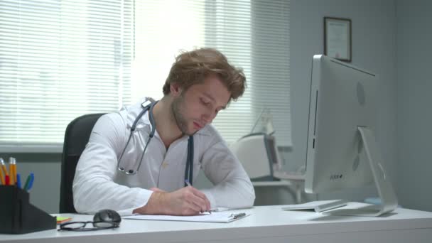 Il dottore si siede a un tavolo in gabinetto di ospedale e scrive — Video Stock