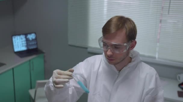 Химик в защитной рабочей одежде принимает трубку с жидкостью и серьезно смотреть в лаборатории — стоковое видео