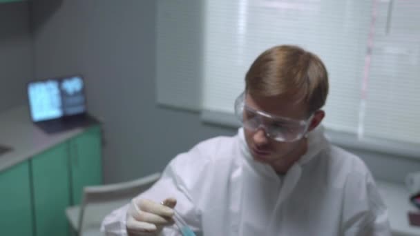 Химик в защитной рабочей одежде принимает трубку с жидкостью и выглядит неодобренным в лаборатории — стоковое видео