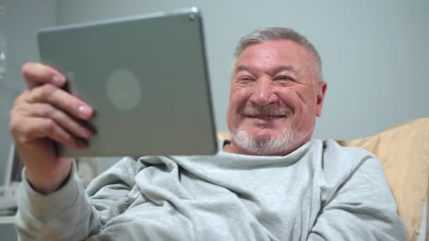 Чоловік лежить на ліжку в лікарні і розмовляє з кимось на планшеті — стокове відео