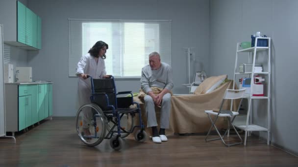 Il medico aiuta il vecchio paziente a sedersi su una sedia a rotelle in ospedale — Video Stock