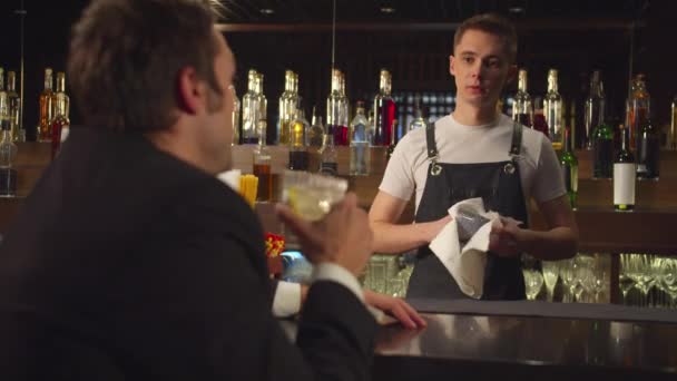 El hombre bebe alcohol y habla con el camarero que limpia un vaso — Vídeo de stock