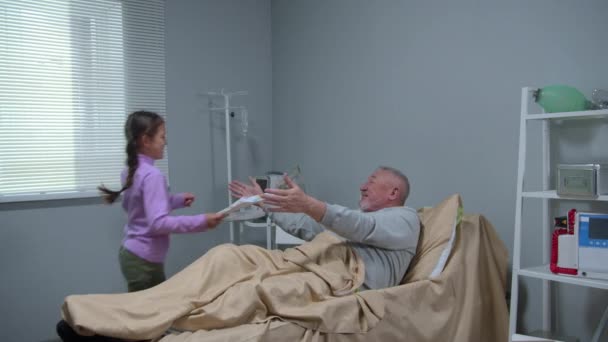 Muž leží v posteli v nemocnici a zdraví svá vnoučata — Stock video