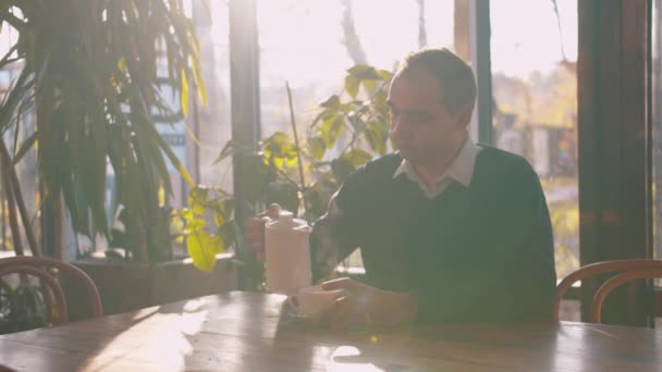 Мужчина сидит в кафе и наливает чай в чашку — стоковое видео