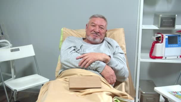 El hombre se acuesta en la cama en el hospital y mira a la cámara con sonrisa — Vídeos de Stock