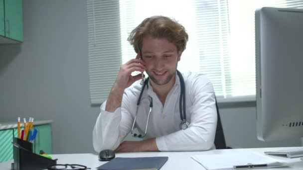 Doctor sit at a table in cabinet of hospital and talk on phone friendly — Stock Video