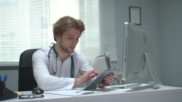 O doutor senta-se em uma mesa no armário do hospital e trabalha com a pastilha — Vídeo de Stock