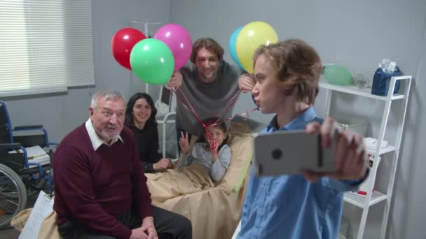 Příbuzní navštíví dívku, která leží na posteli v nemocnici, chlapec udělat skupinovou fotku — Stock video