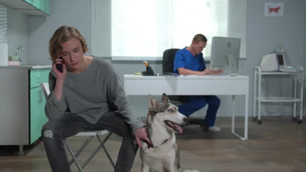 Muž sedí s chraplavý a mluví po telefonu v kabinetu veterináře — Stock video