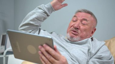 Man lie on the bed in the hospital and talk agressively on tablet