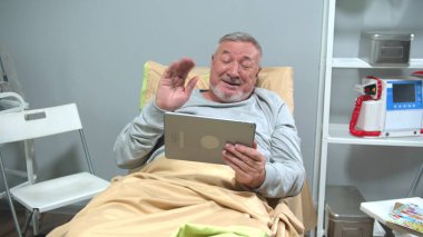 Man lie on bed and talk with someone on the tablet and wave his hand