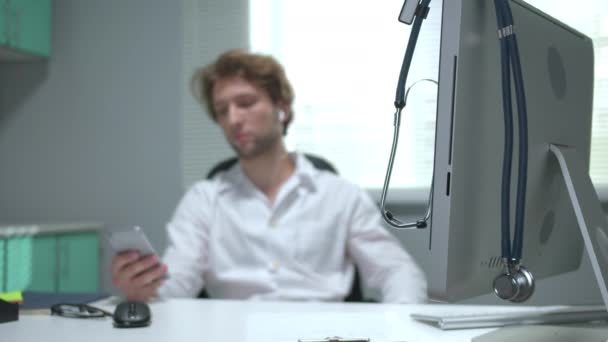 Zwolniony ruch, lekarz siedzi przy stole z telefonem w dłoni na tle stetoskopu — Wideo stockowe