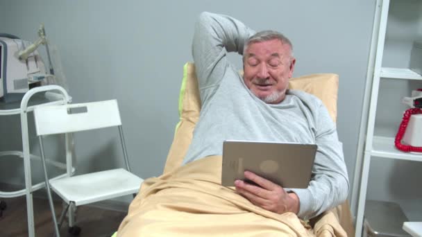 Muž leží na posteli v nemocnici a mluví s někým na desce — Stock video