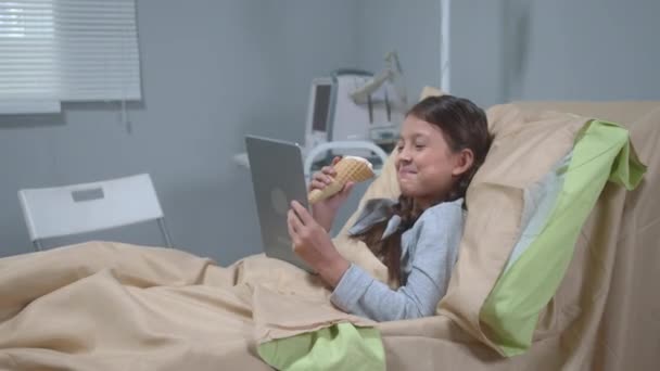 Meisje ligt op bed, eet ijs, kijkt naar iets en lacht op tablet — Stockvideo