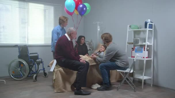 Flicka ligga på sängen på sjukhuset, pojke hålla luft ballonger, andra släktingar prata med henne — Stockvideo