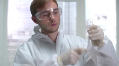 Koruyucu iş elbisesi giyen genç bir bilim adamı kimyasal maddeyle sallanıyor ve laboratuvara giriyor.