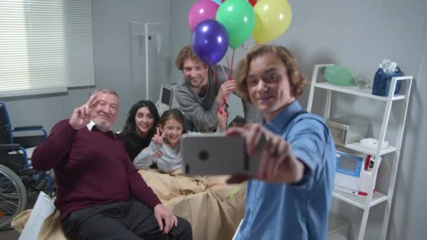 Familieleden bezoeken meisje die op het bed liggen in het ziekenhuis, jongen maakt een grappige groepsfoto — Stockvideo